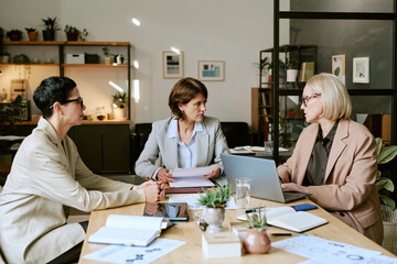 Three middle aged Caucasian women sitting at table discussing documents and working on laptop in...