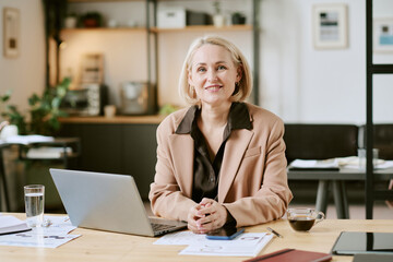 Portrait of middle aged Caucasian woman sitting at desk smiling into camera, working on laptop with...