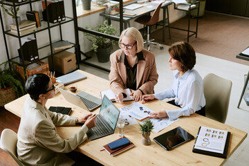 Three middle aged Caucasian women discussing business strategy at table, analyzing financial charts and using laptops in modern office, collaborating on project together