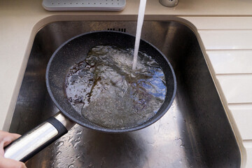 Hand holds frying pan with hot oil under faucet in stainless sink, water hitting oil creates risky splash and steam