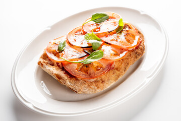 An Italian bruschetta appetizer on a white plate, featuring toasted bread topped with sliced tomatoes, fresh basil leaves, and a glossy balsamic glaze drizzle