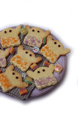 Halloweeen shortcrust cookies in shape of a ghost with white chocolate glaze and colorful sprinkles on a lilac plate on wooden table 