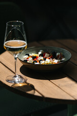 Glass of white wine and bowl of Greek salad with feta, olives, and vegetables on wooden outdoor table in sunlight. Summer lunch, relaxation, Mediterranean lifestyle.
