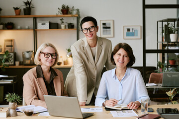 Three Caucasian middle aged women collaborating in modern office, two sitting at desk with laptop...