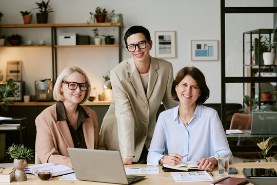 Three Caucasian middle aged women collaborating in modern office, two sitting at desk with laptop and documents, one standing behind smiling, all wearing eyeglasses and business attire