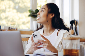 Beautiful smiling African American woman holding mobile phone, looking away, sitting at home working
