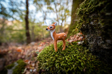 Charming miniature deer figurine stands on lush mossy forest floor, capturing whimsical woodland magic and autumnal enchantment.