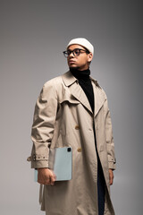 Stylish young man poses confidently with tablet in trendy outfit against a neutral backdrop