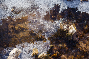 Clear water flows around shimmering ice formations and visible riverbed rocks, capturing a crisp winter or early spring scene