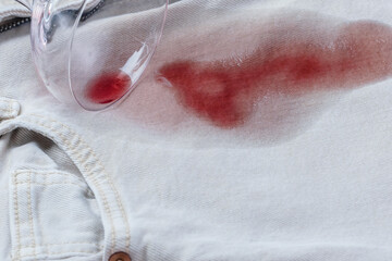 Spilled glass with red wine falled a top a light blue pair of jeans, while a noticeable red stain spreads from the glass. Daily stains concept. top view