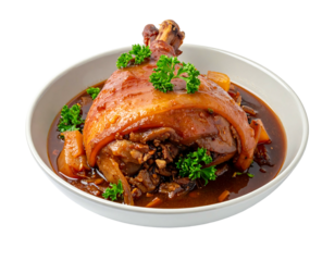 A large, glazed ham hock with gravy, parsley, and veggies served in a white bowl against a transparent background