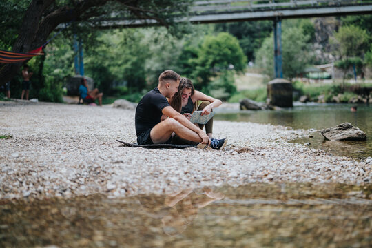 A couple sits on a pebble beach by a river, admiring a map and enjoying nature. The scene captures relaxation, exploration, and the joy of spending time outdoors together.