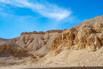 Close up of the steep, barren rock faces and sunbaked mountains of the Valley of the Kings in Luxor Egypt against a deep blue sky with wispy clouds.