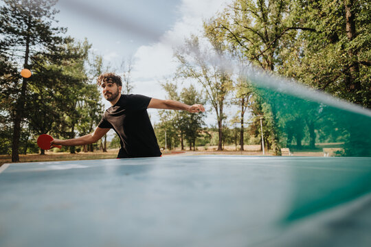 A man in a black shirt hits a ping pong ball on an outdoor table tennis setup in a leafy park. The scene conveys focus, athleticism, and casual outdoor recreation.