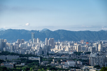 A panoramic view of the city skyline with distant mountains in the background, showcasing modern high-rise buildings and lush greenery under a clear blue sky