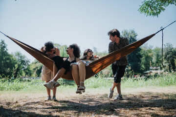 A group of friends shares a playful moment in a hammock set between trees in a park. They smile, chat, and enjoy a warm, sunny day together, embodying leisure and companionship.