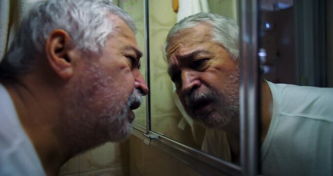 Older man pressing forehead and hand against mirror, deep in thought with a pained expression, capturing a raw and emotional moment of vulnerability and self-reflection