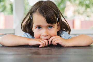 Little girl with dark hair and blue eyes resting her chin on a wooden table, looking curiously at...