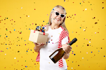 Excited senior woman with gift and bottle of sparkling wine on yellow background with flying...