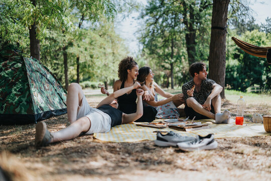 A group of friends relaxes on a checkered blanket under trees, sharing snacks and drinks near a camouflage tent on a sunny day in the forest.