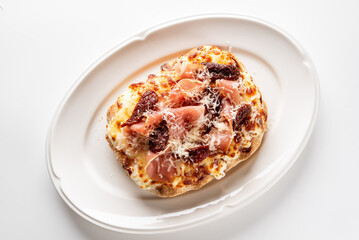 A gourmet toasted bread slice topped with melted cheese, prosciutto, sun-dried tomatoes, and shaved Parmesan on a white plate, photographed on a clean white background.