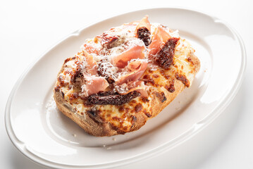 A gourmet toasted bread slice topped with melted cheese, prosciutto, sun-dried tomatoes, and shaved Parmesan on a white plate, photographed on a clean white background.