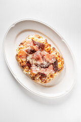 A gourmet toasted bread slice topped with melted cheese, prosciutto, sun-dried tomatoes, and shaved Parmesan on a white plate, photographed on a clean white background.