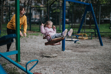 A mother helps her smiling daughter enjoy a swing set in a green park. This cheerful outdoor scene captures the bond of family and the joy of playfulness in a safe environment.