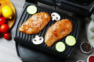 Electric grill with raw marinated chicken fillet, mushrooms, spices and products on wooden table, flat lay
