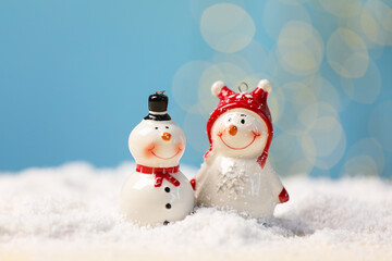 Cute decorative snowman on artificial snow against light blue background with blurred lights, closeup