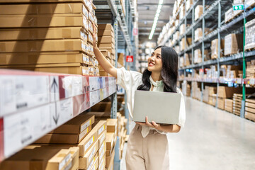 Engaging in meticulous inventory management, a young asian woman professional examines products while using a laptop computer, surrounded by organized shelves filled with boxes