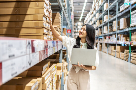 Engaging in meticulous inventory management, a young asian woman professional examines products while using a laptop computer, surrounded by organized shelves filled with boxes