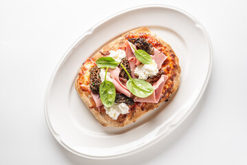 delicious oval flatbread pizza with ricotta cheese, prosciutto, spinach, and morel mushrooms on a white oval plate.