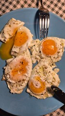 Four sunny-side-up fried eggs seasoned with black pepper on a light blue plate, ready for breakfast. One yolk is broken, with a shiny fork and knife visible on a checkered tablecloth.