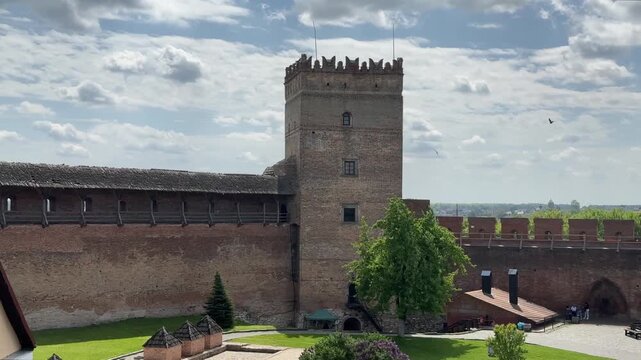 A high-angle view of Lubart's Castle in Lutsk, Ukraine, shows the main tower, brick walls, and the courtyard with green grass, trees, and small buildings under a cloudy sky