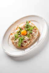 An elegant single serving pizzetta topped with creamy cheese, fresh arugula, shredded crab meat, and a bright raw egg yolk on an oval white plate on a white surface