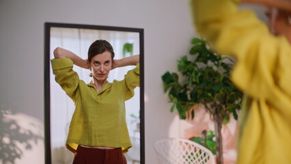Smiling brunette making hairstyle looking mirror at apartment living room.