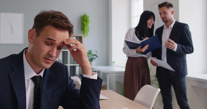 A frustrated office worker sits apart, holding his forehead in stress, while colleagues whisper and discuss him. The workplace scene highlights bullying, gossip, conflict and social isolation.
