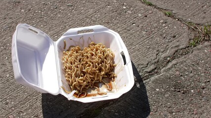 Discarded takeout instant ramen noodles in a white styrofoam container on a gritty outdoor concrete street pavement during a sunny day urban food waste concept