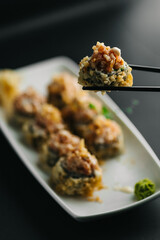 Close-up of deep-fried tempura sushi roll held with chopsticks. Crispy texture, sauce topping, and blurred sushi plate background.
