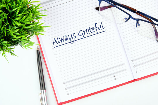 A journal with the words Always Grateful is open on a neat desk. Next to it lies a pen and a pair of glasses, accompanied by a small green plant for a refreshing touch