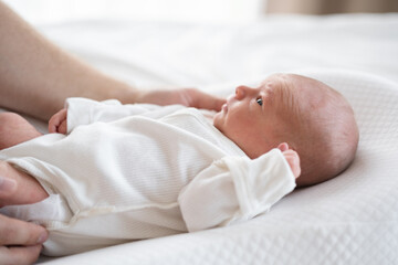 A cute newborn baby sleeps in a white bodysuit on a bed. He lies on a white blanket at home.