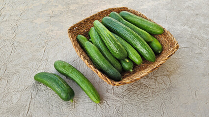 An overhead shot of deep green cucumbers, whole and glossy, piled in a square, rustic wicker basket on a light-colored, wrinkled fabric surface