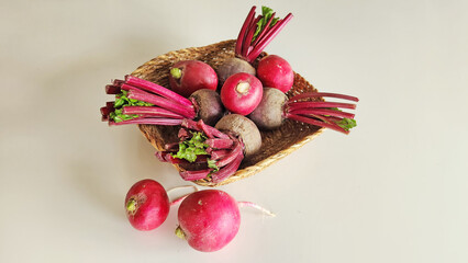 An overhead view of freshly harvested radishes and beets with vibrant burgundy stalks and small green leaves, piled in a rustic square basket on a clean, light-colored surface