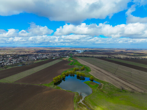 High aerial landscape of vast brown and green agricultural fields, featuring a small reflective pond and a distant village under a dramatic blue, cloudy sky - Powered by Adobe