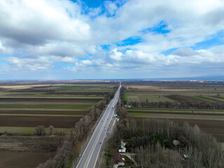 Fototapeta premium High aerial view of a long, straight highway cutting through vast agricultural fields of alternating brown and green strips under a bright, cloudy sky