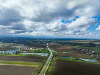 Wide aerial view of a straight canal and several interconnected water bodies running through dark brown and green agricultural fields in a flat valley