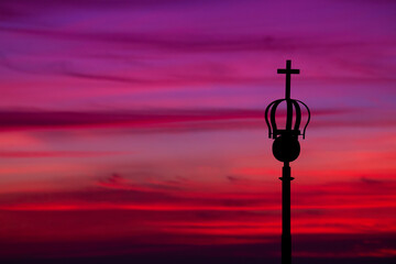 A lamppost crowned with a cross stands against a winter sunset, where deep blues and soft pinks...