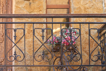 A porch with a metal railing in front of the entrance door of a vintage-style building. A fragment of a stone wall with a flower pot in front of the window. An element of outdoor decor.