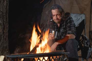A man camping sitting by the fire eating canned food.